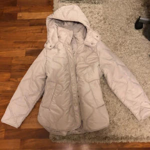 Beige quiltad jacka - Säljer en beige quiltad pufferjacka från H&M med avtagbar huva och snygga vågiga sömmar. Jackan har knappar framtill, två fickor och en lite oversized passform. Perfekt för kyliga dagar och enkel att styla till jeans eller leggings💗💗