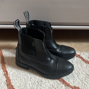 Svarta ridskor från Horze - Snygga svarta ridstövletter från Horze i skinn med dragkedja framtill och dekorativa perforeringar på tån. Klassisk design med låg klack och elastiska paneler på sidorna för extra komfort. Perfekta för dig som gillar stilrena boots till ridning.