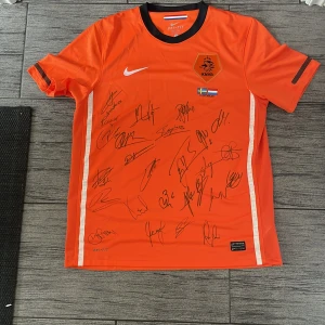 Holland fotbollströja Nike L signerad - Säljer en orange officiell Holland fotbollströja från Nike, storlek L, med svarta detaljer och KNVB-emblem på bröstet. Tröjan har korta ärmar, Dri-FIT-material och är signerad av flera spelare. Matchdetaljer mot Sverige 11 oktober 2011 tryckt på framsidan.
