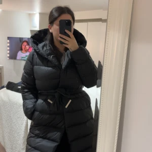 Svart lång dunjacka från BOSS - Säljer en svart lång dunjacka från BOSS med huva och bälte i midjan. Jackan är quiltad och har en snygg, puffig look. Perfekt för kalla dagar och har en stilren design med diskreta detaljer. BOSS-logga på bältet och innerficka.