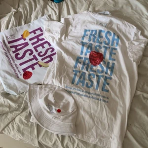 Vit XQS t-shirt med färggrafiskt tryck - Säljer en vit t-shirt från XQS med stort grafiskt tryck på framsidan. Ena t-shirten har blå text 'FRESH TASTE' och ett hallonmotiv, den andra har lila text med hallon, citron och äpple. Båda är kortärmade och gjorda i mjuk bomull. Perfekt för dig som gillar färg och fruktiga vibes.
