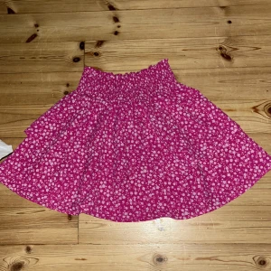 Rosa blommig kortkjol med volang - Supersöt rosa kortkjol med små vita blommor och volangkant. Kjolen har smock i midjan för en bekväm passform och är gjord i ett lätt bomullstyg. Perfekt för varma dagar när du vill känna dig både chill och söt.