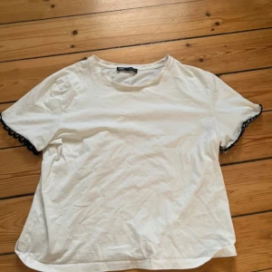 Vit t-shirt med spets från Zara - Vit t-shirt från Zara i mjuk bomull med rund hals och korta ärmar. Ärmsluten har svart spetsdetalj som ger en cool kontrast. Passformen är normal och modellen är klassisk med en liten twist. Perfekt för dig som gillar enkla plagg med en unik detalj.