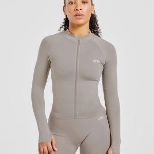 EMPOWER SEAMLESS FULL ZIP JACKET taupe  - Slimmad grå zip-tröja från AYBL med lång ärm och hög krage. Tröjan har dragkedja framtill och är perfekt för träning. Snygg och enkel design med diskret logga på bröstet. Aldrig använd ser ut som på bilden