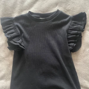 Svart volangtopp från Gina Tricot - Supersnygg svart topp från Gina Tricot med ribbad struktur och volangdetaljer på axlarna. Toppen är ärmlös och har en rund halsringning. Perfekt att styla med jeans eller kjol för en trendig look. Den är köpt 2022 men inte använd sen dess. Den är fräsch utan fläckar. Säljer för 80kr men pris kan diskuteras för den är gammal osv. Köp inte på ”KÖP NU”, betalningen genomförs i chatten med Swish. Frakten kan bli runt 30-50kr. 