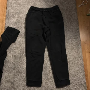 Svarta Nike tech byxor - Svarta mjukisbyxor från Nike i storlek L. Byxorna har dragsko i midjan, muddar vid bensluten och en dragkedjeficka på sidan. Perfekta för chill eller träning, med klassisk Nike-logga och mjukt material.