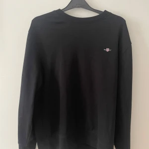 Svart sweatshirt från Gant - En stilren svart sweatshirt från Gant med liten broderad logga på bröstet. Klassisk rund halsringning och långa ärmar. Perfekt för dig som gillar enkel och tidlös stil.