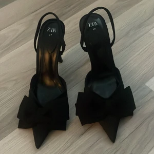 Svarta pumps med rosett från Zara - Stilrena svarta pumps från Zara med spetsig tå och hög klack. Skorna har en elegant rosett framtill och smal rem runt hälen. Perfekta för dig som vill sticka ut med en klassisk men trendig look.