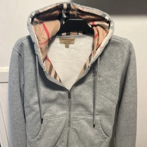 Grå hoodie från Burberry med mönstrad huva - Säljer en grå hoodie från Burberry med dragkedja och klassisk rutig huva i beige, svart och rött. Tröjan har snörning i huvan, två fickor fram och är långärmad. Perfekt för dig som gillar stilrena plagg med en twist av lyx.
