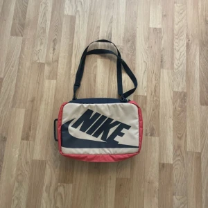 Nike axelväska i beige och svart - Säljer en stilren axelväska från Nike i beige och svart med röda detaljer. Väskan har stort Nike-tryck på framsidan, justerbar axelrem och praktiskt bärhandtag. Invändigt finns ett stort fack och meshficka med dragkedja. Perfekt för att bära med sig det viktigaste.