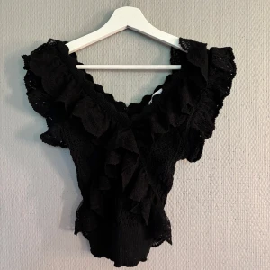 Svart spetsblus med volanger från Zara - Supersnygg svart blus från Zara i storlek S, med spetsdetaljer och stora volanger längs v-ringningen och axlarna. Blusen har korta ärmar och är croppad med ribbad nederkant. Perfekt för dig som gillar statement-plagg och coola detaljer.