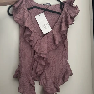 Lila volangblus från Zara - Supersöt lila blus från Zara med volanger längs v-ringningen och ärmsluten. Blusen har en skrynklig struktur och omlottkänsla framtill. Perfekt för dig som gillar romantiska detaljer och vill sticka ut lite extra.
