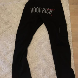 Svarta mjukisbyxor från Hoodrich - Svarta mjukisbyxor från Hoodrich med stor logga framtill och mindre tryck bak. Byxorna har dragsko i midjan och en ficka på sidan. Sköna och stilrena, perfekta för chill eller streetwear. Materialet är mjukt och bekvämt.