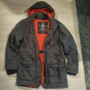 Svart parkajacka från Hampton Republic - Säljer en svart parkajacka från Hampton Republic med orangefärgat foder och huva. Jackan har flera fickor med både knappar och dragkedja, samt ribbade muddar vid ärmsluten. Perfekt för kyliga dagar och har en snygg, urban look.