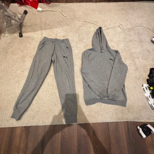 Grå mjukisbyxor från Puma - Snygga grå mjukisbyxor från Puma med elastisk midja och ribbade muddar vid bensluten. Byxorna har två sidofickor och en svart Puma-logga på benet. Perfekta för chill eller träning, tillverkade i mjukt bomullsmaterial för maxad komfort.