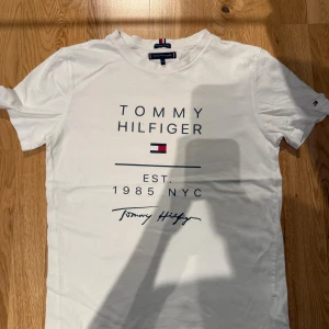 Vit Tommy Hilfiger t-shirt i bomull - Vit t-shirt från Tommy Hilfiger med tryck på bröstet och logotyp i klassiska färger. Rund hals och korta ärmar. Tillverkad i mjuk ekologisk bomull som känns skön mot huden. Perfekt för dig som gillar stilren design och ikoniska märken.