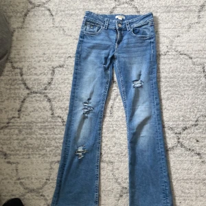 Blå bootcut jeans med slitningar - Snygga blå bootcut jeans med slitna detaljer på benen och broderade bakfickor. Jeansen har femficksdesign, knapp och dragkedja framtill samt slitningar
