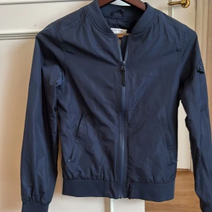 Mörkblå bomberjacka från WearColour - Snygg mörkblå bomberjacka från WearColour med klassisk ribbad krage och muddar. Jackan har dragkedja framtill, två fickor med dragkedja och ett stilrent, minimalistiskt utseende. Perfekt för dig som gillar enkel och clean stil. Osäker på om den är till man eller dam så satte unisex 