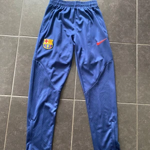 Blå FC Barcelona träningsbyxor Nike - Snygga blå träningsbyxor från Nike med FC Barcelona-logga och röd swoosh på ena benet. Byxorna har elastisk midja och smal passform, perfekta för fotbollsträning eller chill. Tillverkade i polyester för maximal komfort och rörelsefrihet.
