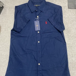 Blå kortärmad skjorta från Ralph Lauren - Snygg marinblå skjorta från Ralph Lauren med korta ärmar och klassisk krage. Skjortan har knappar framtill och den ikoniska röda broderade loggan på bröstet. Tillverkad i bomull, perfekt för en clean och stilren look.