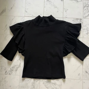 Svart volangtopp från Zara - Svart ribbad topp från Zara med höghalsad krage och unika volangdetaljer längs axlarna. Toppen har trekvartsärmar och är figurnära i modellen. Perfekt för dig som vill sticka ut med en cool och trendig look.
