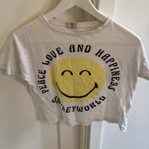 Vit croppad t-shirt SmileyWorld - Vit croppad t-shirt från SmileyWorld med stort gult smileytryck och texten 'Peace Love and Happiness' på framsidan. Rund hals och korta ärmar. Mjuk bomull som är skön mot huden, perfekt för chill dagar. Passar dig som gillar positiva vibes och streetstyle.
