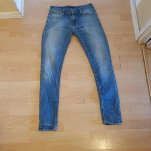 Blå Slim jeans från Nudie Jeans - Säljer ett par klassiska blå Slim jeans från Nudie Jeans med snygga slitningar och orangea sömmar. Jeansen har fem fickor och en smal passform som sitter tajt längs benen. Perfekta för dig som gillar en modern och stilren look.