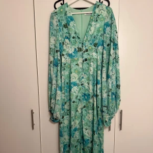 Blommig långklänning från Adoore - Säljer en superfin långklänning från Adoore med blommigt mönster i gröna och blå toner. Klänningen har v-ringning, långa puffärmar och knappar framtill. Perfekt för dig som vill ha en flowy och färgglad look.
