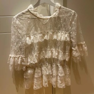 Spetsblus - Superfin blus i ljus spets med volanger och genombrutet mönster. Blusen har trekvartsärm och hög hals med volangdetaljer. Perfekt för dig som gillar en romantisk och feminin stil.