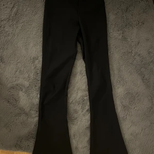 Svarta bootcut kostym byxor med ficka - Snygga svarta kostym byxor med bootcut passform och en ficka med knapp bak. Byxorna har hög midja och är perfekta för dig som gillar en klassisk men trendig look. Materialet känns mjukt och följsamt, och de har bälteshällor i midjan.