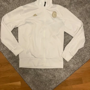 Vit Adidas AC Milan track jacket - Vit track jacket från Adidas med AC Milan-logga i guld på bröstet. Jackan har hel dragkedja, ståkrage och två fickor med dragkedja. Materialet är mjukt och sportigt, perfekt för fotbollsfans som vill ha en clean och stilren look.