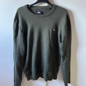 Olivgrön stickad tröja från WE - Säljer en olivgrön stickad tröja från WE med rund hals och lång ärm. Tröjan har en diskret gul broderad detalj på bröstet och en grovstickad struktur som ger en cool känsla. Perfekt för lager-på-lager under hösten och vintern.