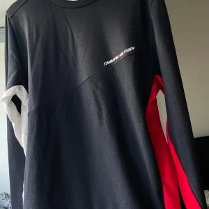 Tommy Hilfiger långärmad tröja M - Snygg långärmad tröja från Tommy Hilfiger i svart med röda och vita detaljer på ärmarna. Klassisk logga på bröstet och rund halsringning. Perfekt för dig som gillar streetwear och vill sticka ut med stilrena färgblock. Materialet känns mjukt och bekvämt.