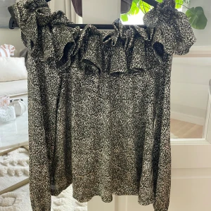 Glittrig off-shoulder blus från Ida Sjöstedt - Supersnygg blus från Ida Sjöstedt i glittrigt svart och silvermönster. Klänningen har volangdetaljer upptill och är tillverkad i ett skimrande material som verkligen sticker ut. Perfekt för dig som vill ha något extra till festen!