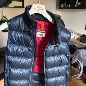 Mörkblå dunväst Parajumpers Sheen XS - Snygg mörkblå dunväst från Parajumpers, modell Sheen. Västen har quiltad design, hög krage och dragkedja framtill. Insidan är röd för en cool kontrast. Perfekt att slänga över en hoodie för extra värme och stil. Kommer med original klädhängare från parajumpers. 