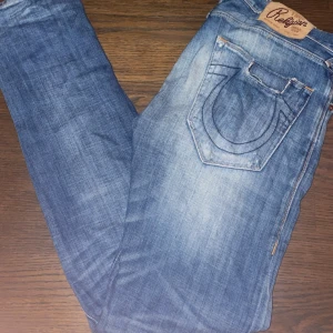True Religion blå jeans med broderad ficka - Säljer ett par blå jeans från True Religion med ikonisk broderad bakficka och slitna detaljer. Jeansen är tillverkade i 100% bomull och har klassisk femficksdesign, kopparfärgade knappar och kontrastsömmar. Perfekta för dig som gillar streetwear och vill sticka ut.