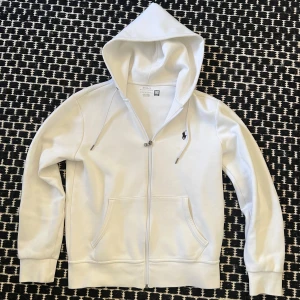 Vit hoodie från Ralph Lauren - Säljer en stilren vit hoodie från Ralph Lauren med dragkedja och klassisk logga på bröstet. Perfekt för en avslappnad look. Den har långa ärmar och en praktisk huva med snören.