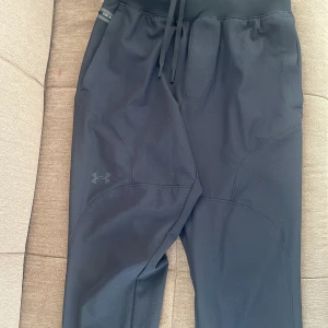 Svarta träningsbyxor från Under Armour - Snygga svarta träningsbyxor från Under Armour med elastisk midja och snörning. Perfekta för träning med en bekväm passform och stilren design. Byxorna har en diskret logga på benet. lägg ett pris själv 
