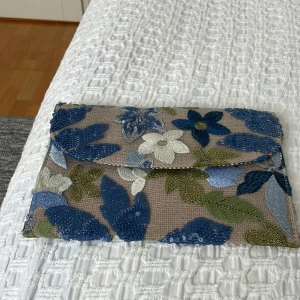 Blommig kuvertväska med paljetter - Säljer en snygg kuvertväska med blommigt mönster i blått, grönt och vitt. Väskan har paljett-detaljer och ett randigt foder inuti. Perfekt för att ge en färgklick till din outfit!