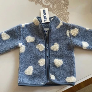 Blå fleecejacka - Säljer en söt blå fleecejacka från RIKKIK med vita hjärtan. Jackan har en dragkedja framtill och är långärmad. Perfekt för att hålla sig varm och mysig. Storlek 6-9 månader💕