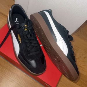 Svarta sneakers från Puma - Snygga svarta sneakers från Puma med vita detaljer och en brun sula. Skorna har snörning och en klassisk design med guldiga logotyper på sidan och tungan. Perfekta för en stilren look.