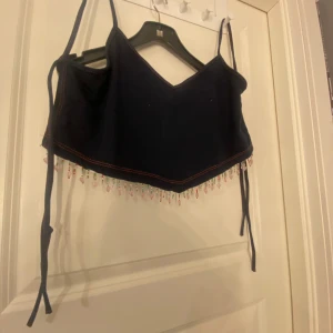 Svart linnetopp med pärldetaljer - Snygg mörkblå linnetopp med smala axelband och v-ringning. Toppen har dekorativa pärlor i olika färger längs nederkanten som ger en unik touch. Perfekt för sommarens alla tillfällen!