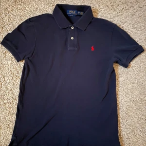 Mörkblå pikétröja från Ralph Lauren - Säljer en stilren mörkblå pikétröja från Ralph Lauren med klassisk krage och två knappar. Tröjan har en röd broderad logotyp på bröstet och är i slim fit. Perfekt för en avslappnad men ändå elegant look.