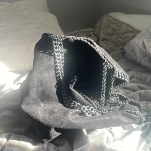 Svart axelväska med kedjedetaljer - Snygg svart axelväska i mocka med kedjedetaljer. Perfekt för att ge en edgy touch till din outfit. Väskan har en rymlig insida och stilrena kedjehandtag.