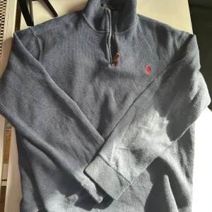 1/3 zipper Polo ralph lauren tröja  - Säljer en stilren blå tröja från Polo Ralph Lauren med halv dragkedja och röd broderad logga på bröstet. Perfekt för en avslappnad men snygg look. Tröjan har långa ärmar och är gjord i ett mjukt material.