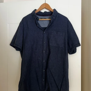 Mörkblå linneskjorta från Dressmann - Säljer en stilren mörkblå skjorta från Dressmann i en skön linneblandning. Skjortan har korta ärmar och knappar framtill samt en bröstficka. Perfekt för en avslappnad men ändå välklädd look. 🌟
