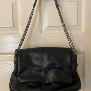 Svart axelväska med kedjedetaljer - Snygg svart axelväska i skinn med kedjedetaljer på axelremmen. Väskan har en stilren design med en praktisk dragkedja upptill. Perfekt för att bära dina dagliga nödvändigheter med stil.