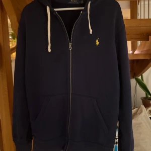 Mörkblå hoodie från Ralph Lauren - Säljer en snygg mörkblå hoodie från Ralph Lauren med dragkedja och vit snörning i huvan. Den har en klassisk gul logga på bröstet och praktiska fickor framtill. Perfekt för en avslappnad stil. Tröjan är lätt lätt solblekt framefrån, dock nästan inte märkbart 
