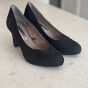 Svarta mockapumps från Tamaris - Snygga svarta pumps från Tamaris i mjuk mocka. De har en klassisk design med en bekväm klack och rundad tå. Perfekta för en elegant look.