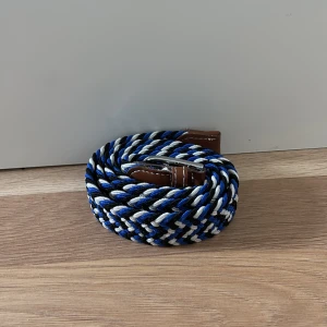 Flätat bälte i blått, vitt och svart  - Snyggt flätat bälte i blått, vitt, svart och brunt med silverfärgat spänne. Perfekt för att ge en färgklick till din outfit. Bältet har bruna skinnliknande detaljer vid spännet.